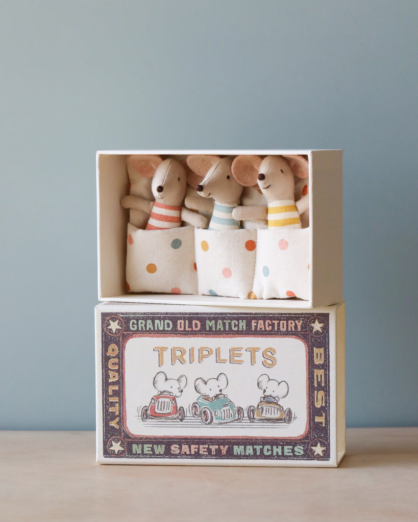 Three Maileg Triplets Baby Mice In Matchbox dressed cute peeking out from a vintage-style matchbox labeled "grand old match factory best triplets new safety matches," set against a soft blue background.