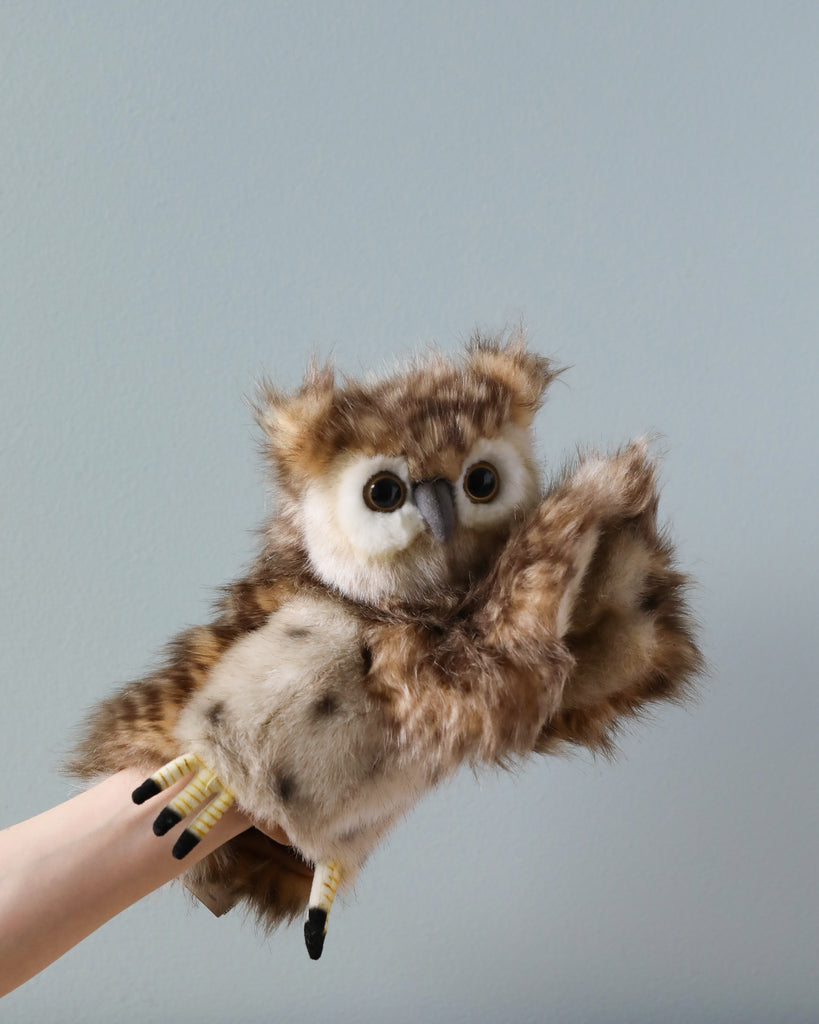 Sentence with product name: A person's hand holding the Owl Puppet, a cute and fluffy brown and white owl puppet against a plain grey background. The realistic stuffed animal has large, round eyes and appears lifelike.