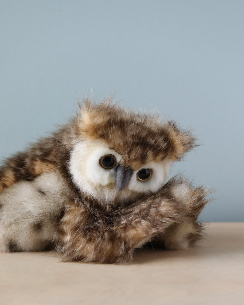 A small fluffy owl puppet with a round face, big yellow eyes, and speckled brown feathers sits hunched on a wooden surface against a pale blue background. This realistic stuffed animal from HANSA