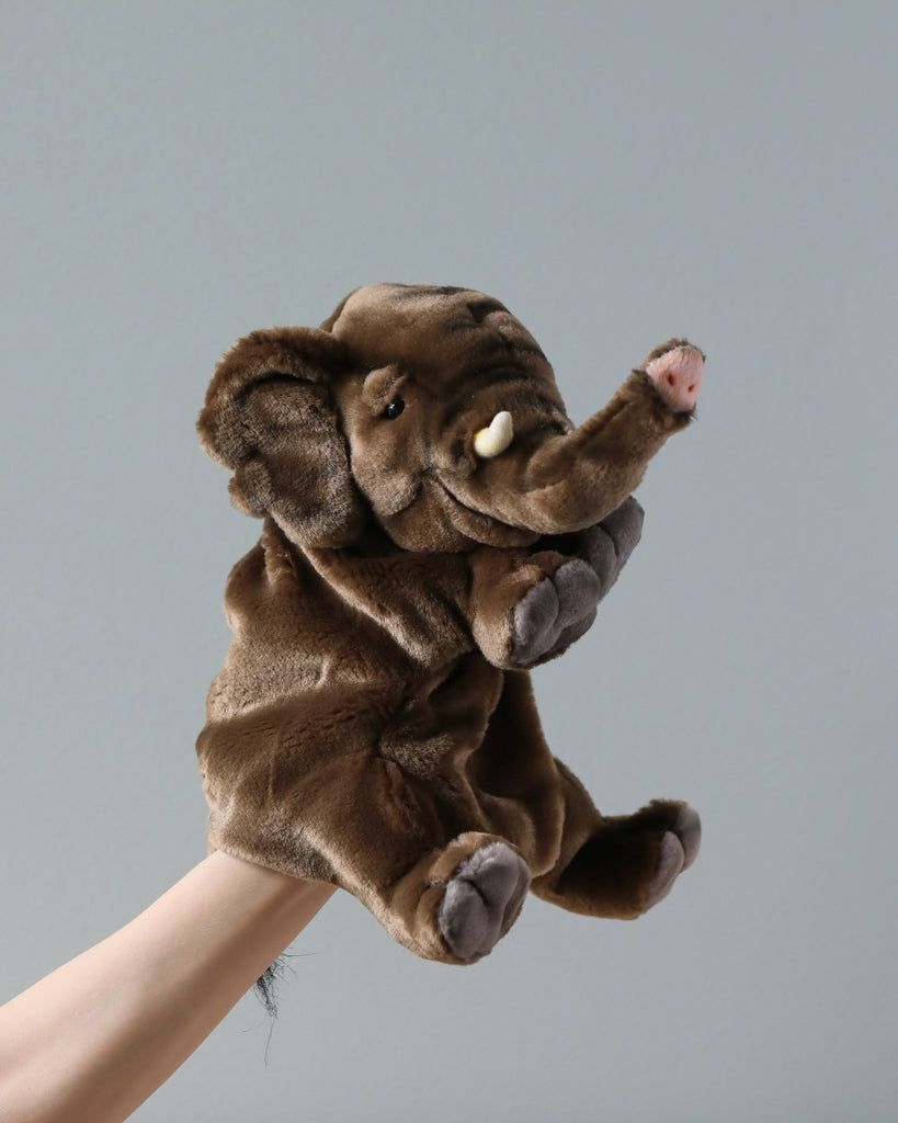 A Elephant Puppet, crafted from quality man-made materials with a raised trunk and floppy ears, is playfully perched on a human hand against a plain gray background.
