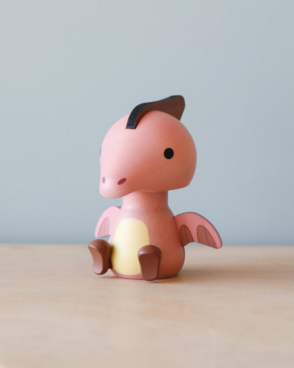 A small, pink Wooden Pterosaur Dinosaur Bobblehead with a darker pink belly, sitting on a wooden surface against a blue background. The toy has a friendly appearance, with big black eyes and a head that shakes