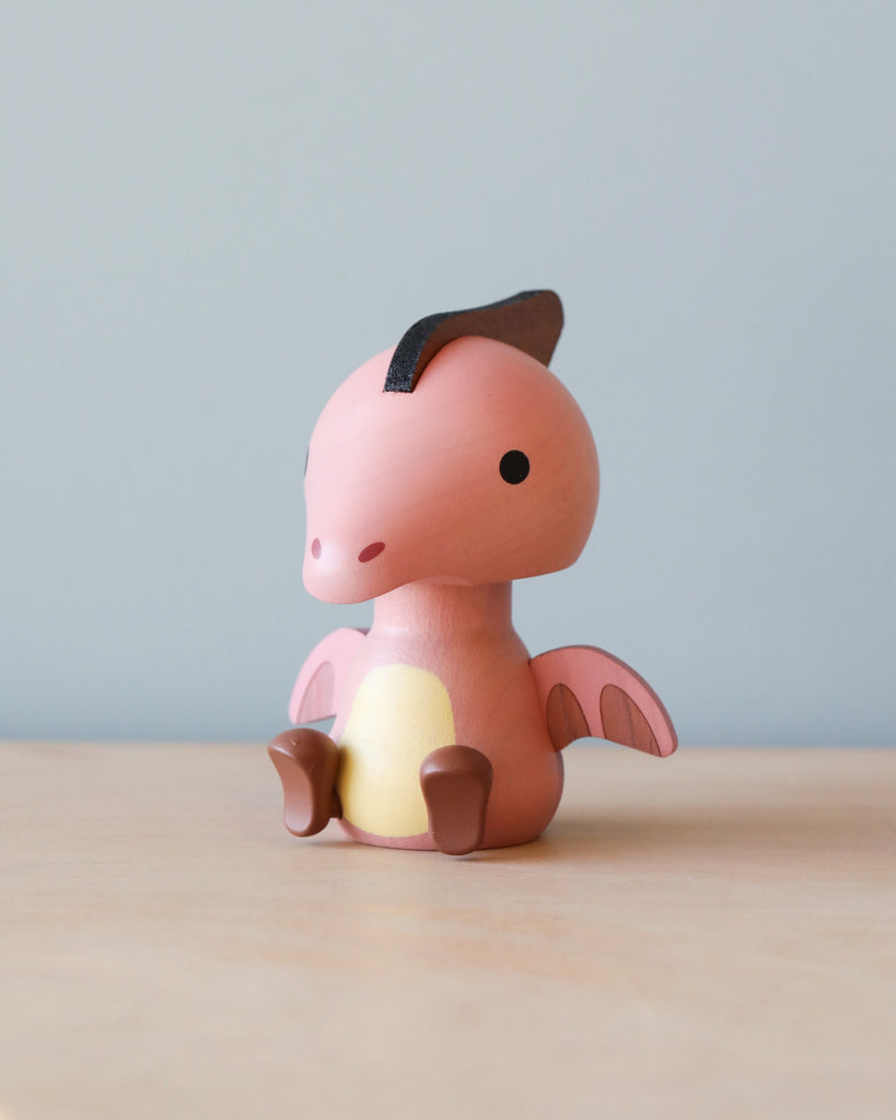 A small, pink Wooden Pterosaur Dinosaur Bobblehead with a darker pink belly, sitting on a wooden surface against a blue background. The toy has a friendly appearance, with big black eyes and a head that shakes