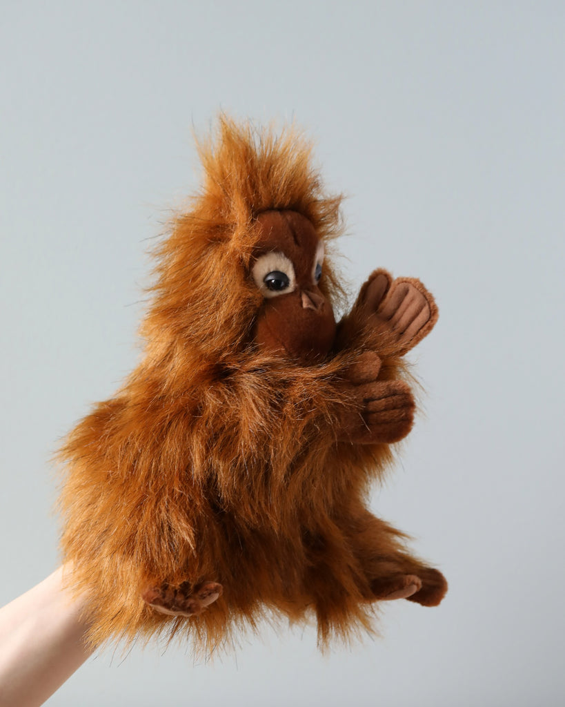 A HANSA Orangutan Puppet with fluffy brown fur is held up against a light gray background, covering its face with one hand in a playful gesture.