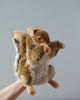 A realistic HANSA Squirrel Puppet is playfully perched on a human finger, depicted against a soft gray backdrop. The puppet mimics a dynamic pose with its tail fluffed and arms open.