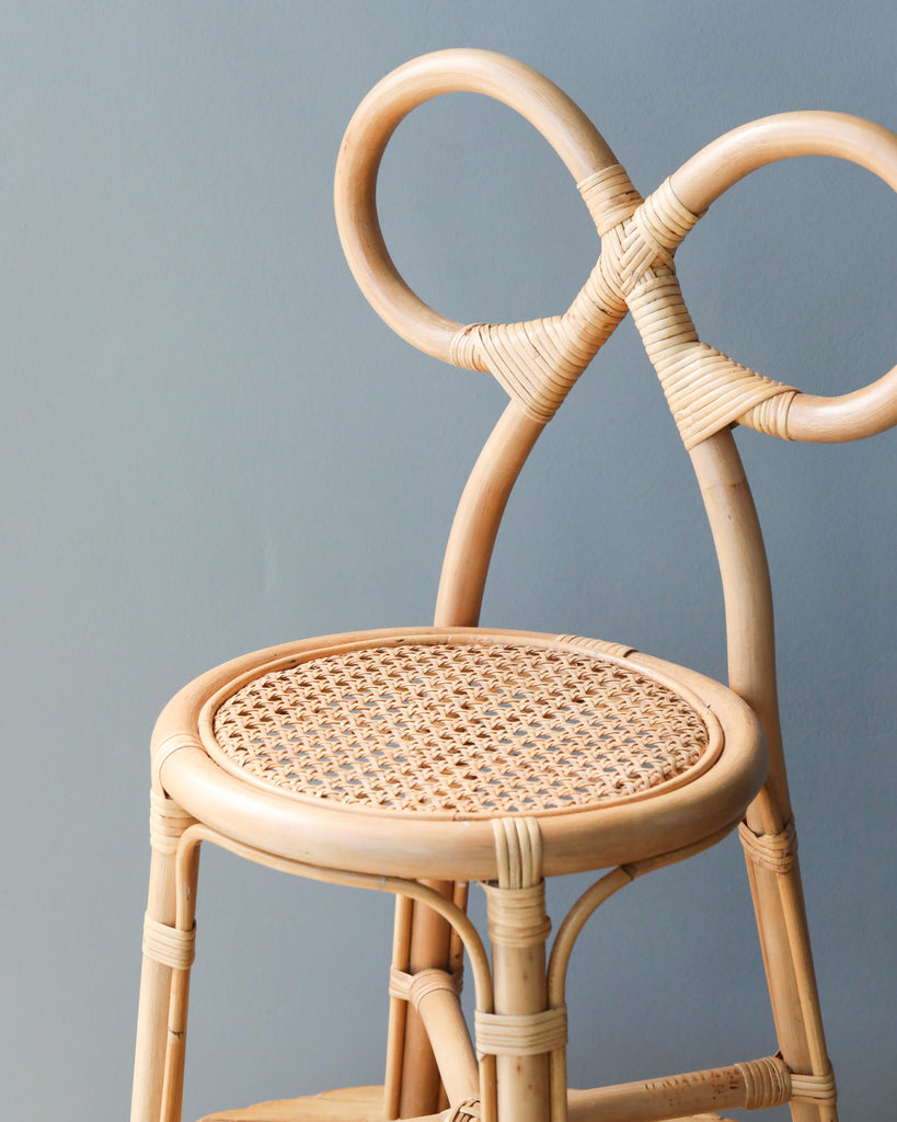 A close-up of a Toddler Rattan Bow Chair with an intricately designed backrest featuring intertwined loops, set against a soft gray background. The chair has a woven seat and light wooden frame.