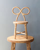 A unique Toddler Rattan Bow Chair with an artistic backrest shaped like a pretzel, placed on a rustic wooden round table against a plain grey background.