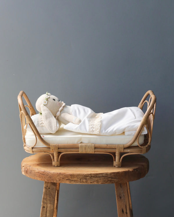 A Poppie Rattan Doll Daybed + Duvet Set lying on a white blanket in a small rattan day bed, which is placed on a rustic wooden stool against a grey background.