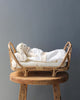 A Poppie Rattan Doll Daybed + Duvet Set lying on a white blanket in a small rattan day bed, which is placed on a rustic wooden stool against a grey background.