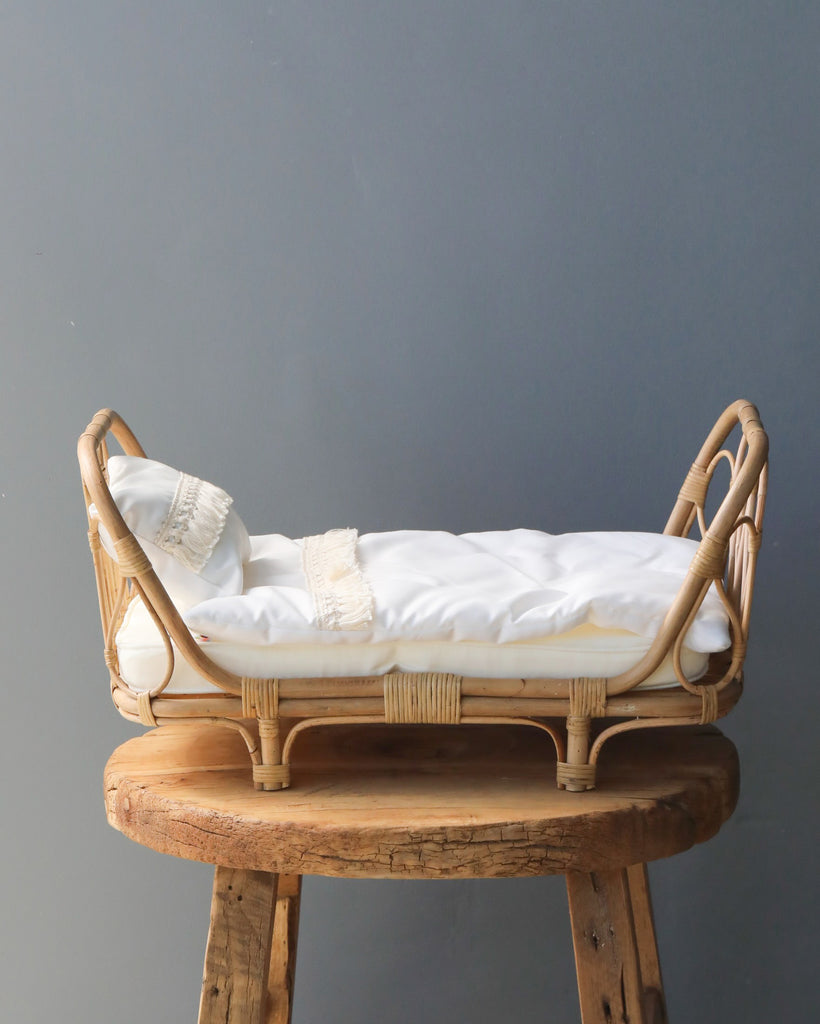 A small Poppie Rattan Doll day bed with white bedding on a rustic wooden stool against a gray background.