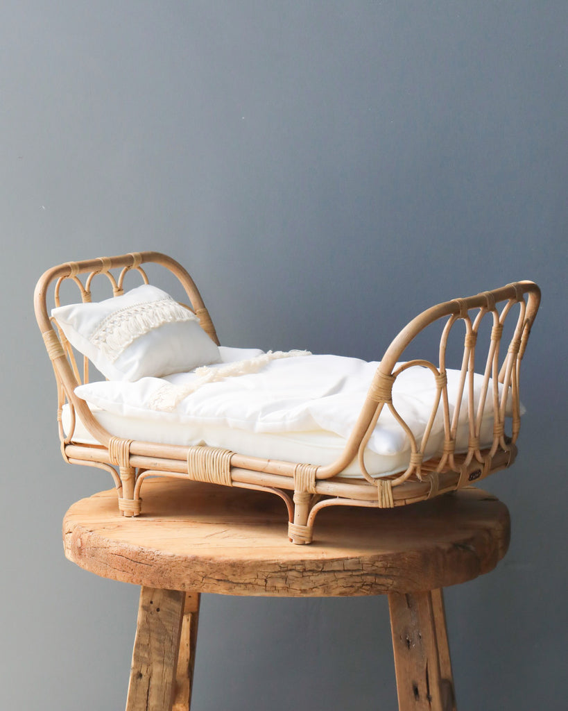 A Poppie Rattan Doll Daybed with white bedding and pillows, displayed on a rustic wooden stool against a plain grey background.
