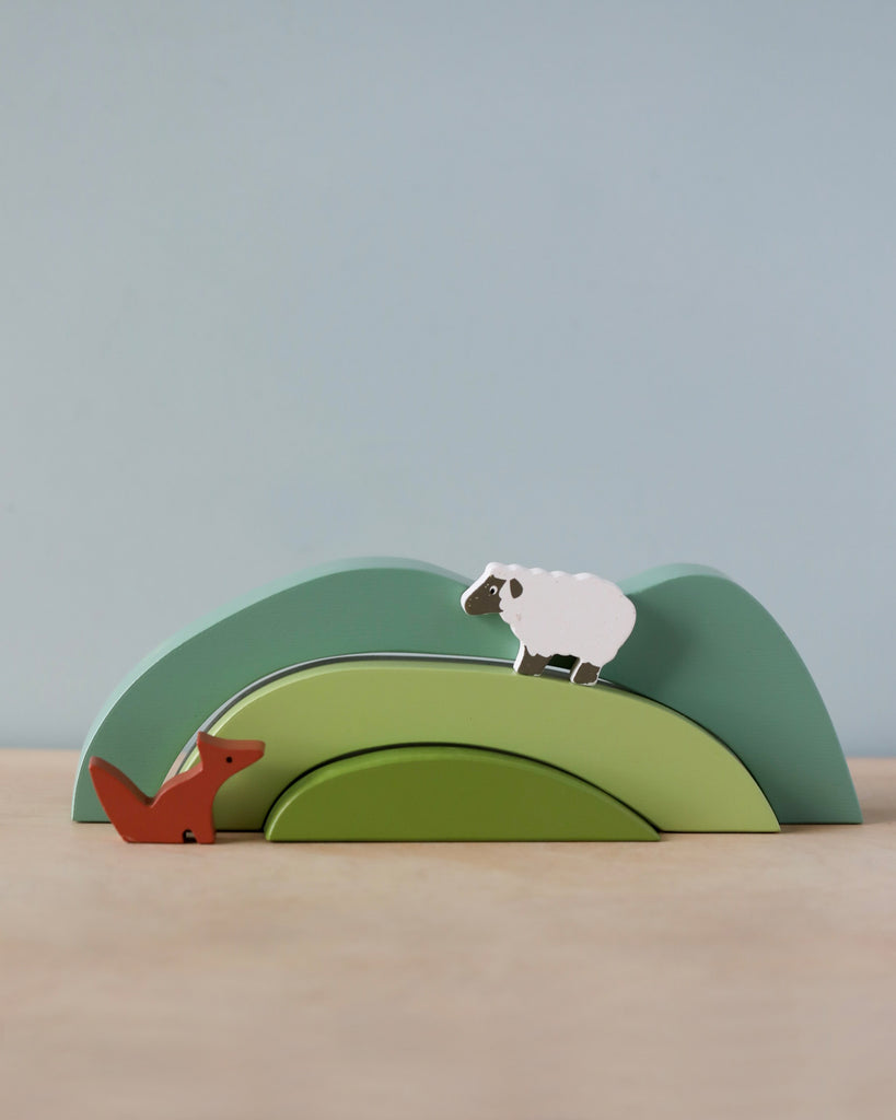 A Green Hill Stacker comprising three nested solid wood hills, forming a hill, with a toy sheep on the top arch and a toy fox on the bottom arch against a pale blue background.