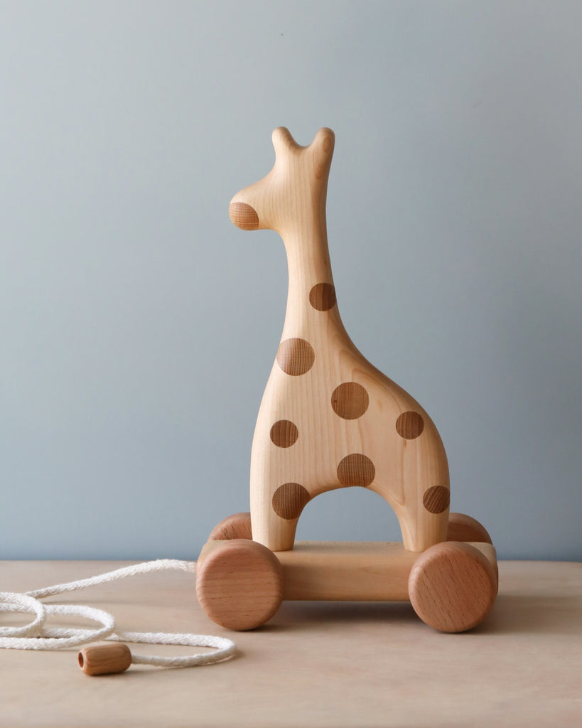 A Handmade Wooden Giraffe Pull Toy with brown spots and four wheels offers endless fun. It features a natural wood finish and a white pull string with an attached handle. The background is simple, showcasing a light blue wall and a light-colored wooden surface—perfect for charming kids' toys collections.