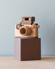 A Father’s Factory vintage style wooden toy camera with a large lens and viewfinder, sitting on top of a small brown block against a pale blue background.