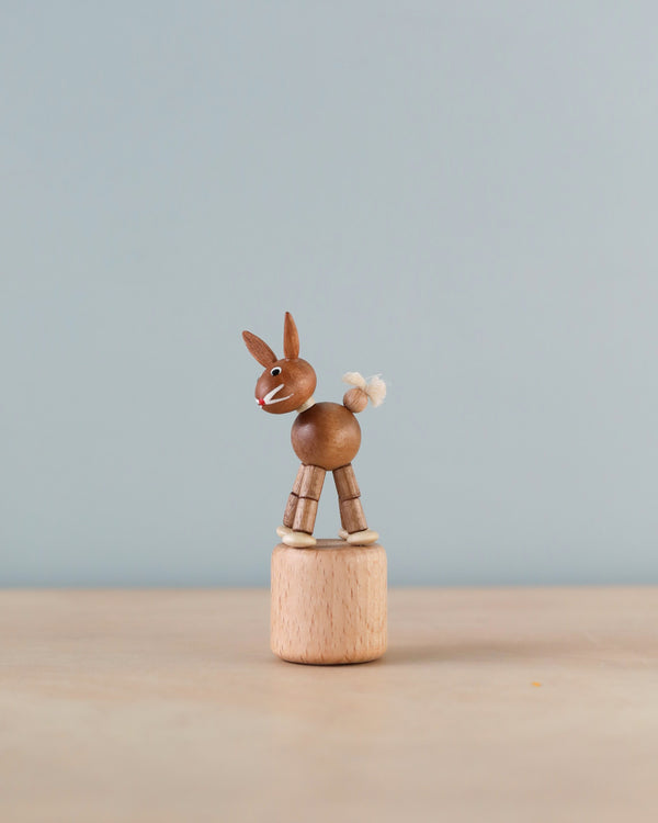 A small wooden figurine of a brown donkey, handmade in Germany, with a white tufted tail, standing on a cylindrical wooden block against a light blue background.