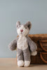 A Senger Naturwelt Stuffed Animal - Cat with gray and white fur, handmade in Germany, sitting upright in front of a woven basket on a wooden table against a pale blue background.