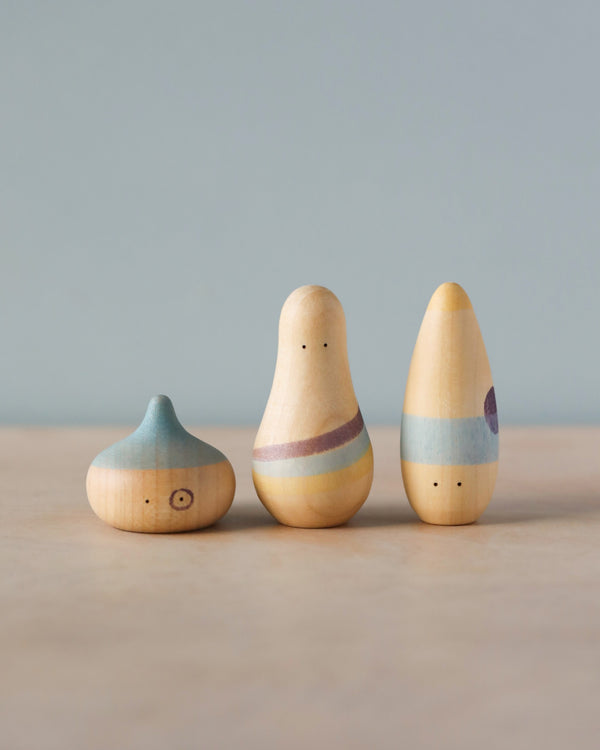 Three whimsical wooden Grapat Wow! Play Set toys resembling people, decorated in pastel colors, arranged on a soft beige background.