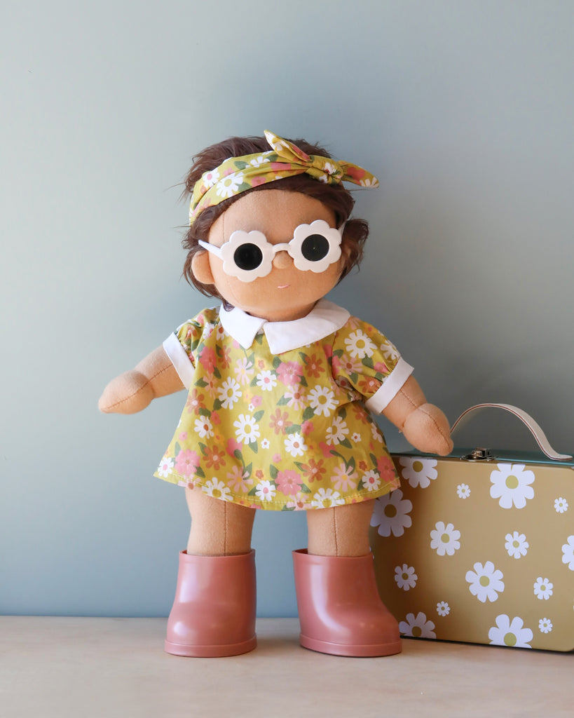 A Dinkum Doll - Sprout (Extended Pack) stands against a light blue background, wearing a yellow dress with white and pink flowers, a matching headband, and white sunglasses. The doll has short brown hair and pink boots. Beside it is a yellow suitcase decorated with white flowers, perfect for any Pretend Pack adventure.