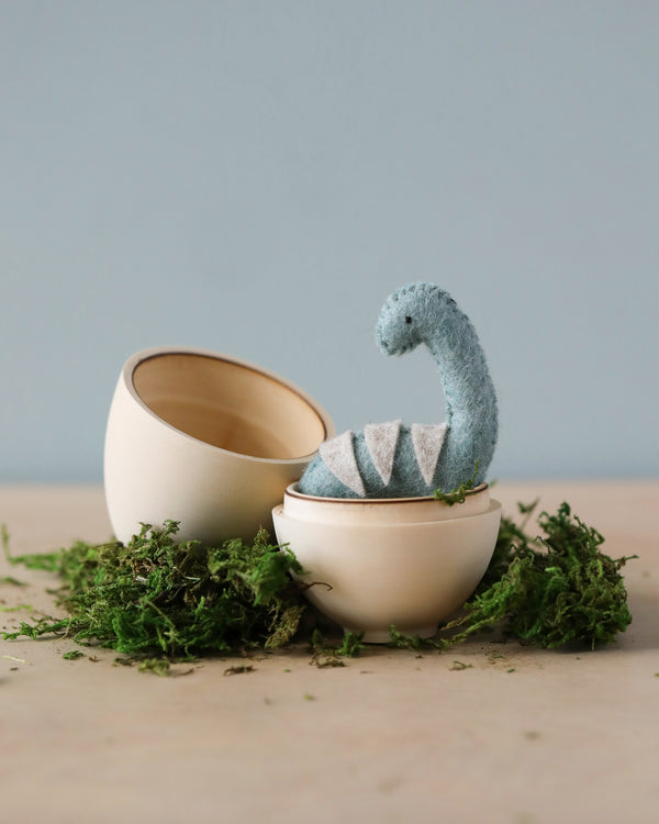 A felted dinosaur toy resting inside a cracked-open Handmade Dinosaur Egg, surrounded by green moss on a wooden surface against a pale blue background.