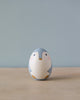 A handcrafted Hand Painted Hollow Wooden Easter Egg - Penguin painted to resemble a cute bird with a grey head, white and pink cheeks, and a simple beige body, set against a soft blue background.