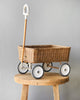 A handmade Olli Ella Rattan Wonder Wagon on wheels with a handle, placed on a round wooden stool against a plain gray background.