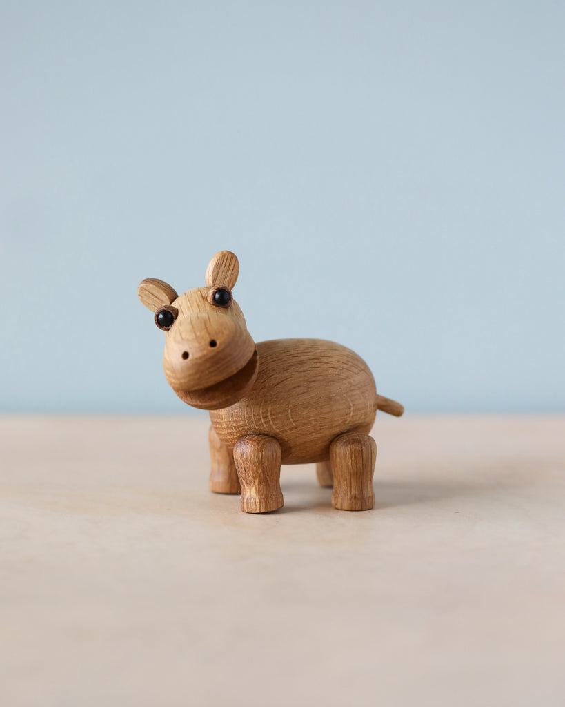 Spring Copenhagen Wilma with a smooth, polished finish. The hippo has a round body, four short legs, a small tail, and black bead-like eyes. Crafted from FSC Oak, it stands on a flat, light-colored surface against a pale blue background, evoking the charm of African wildlife.