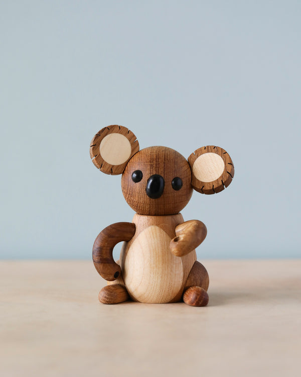 A Spring Copenhagen Matilda wooden koala toy with prominent ears and black eyes sits against a light blue background. Crafted from FSC Oak, the baby koala girl has a two-tone body with darker patches on its.