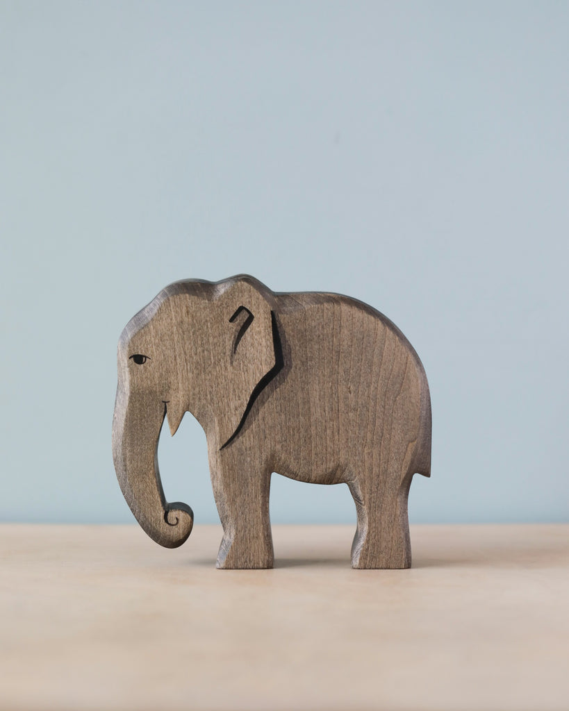 A Handmade Holzwald Elephant figurine, intricately carved with visible grain patterns, stands on a flat surface against a soft blue background. This piece is a fine example of sustainable toys.