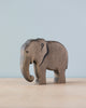 A Handmade Holzwald Elephant figurine with visible wood grain, standing on a plain surface against a soft blue background. The elephant is stylized with a dark, etched mark on its side and crafted from sustainable wood.