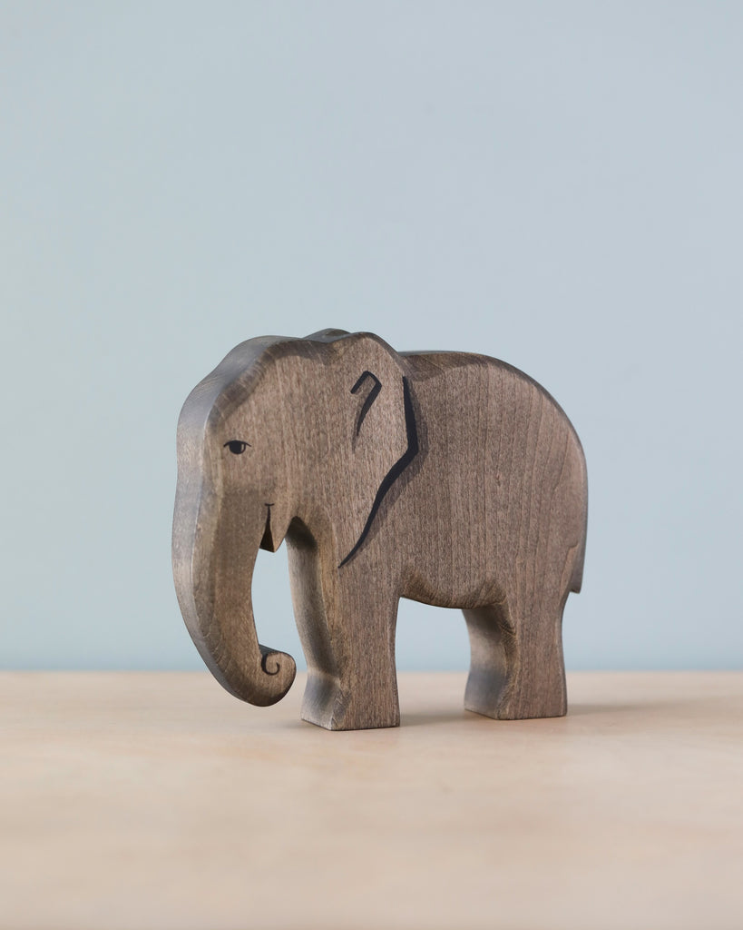 A Handmade Holzwald Elephant figurine with visible wood grain, standing on a plain surface against a soft blue background. The elephant is stylized with a dark, etched mark on its side and crafted from sustainable wood.