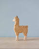 A Handmade Holzwald Alpaca toy stands against a soft blue background. The alpaca, crafted from sustainable wood, is intricately carved with visible wood grain and a lighter color for its face and legs.