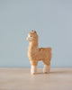 A small, Handmade Holzwald Alpaca stands on a table against a soft, light blue background, showcasing intricate detailing and natural wood grain. This sustainable toy reflects meticulous craftsmanship and environmental consciousness.
