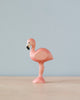 A Handmade Holzwald Flamingo figurine, painted pink, stands on one leg with a plain, light blue background.