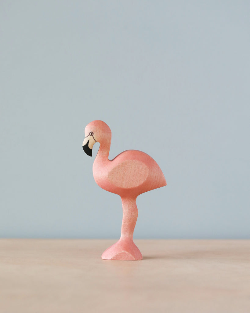 A Handmade Holzwald Flamingo figurine, painted pink, stands on one leg with a plain, light blue background.