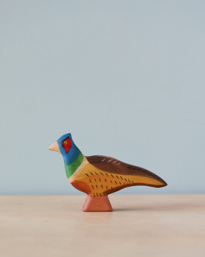 A colorful sustainable handmade Holzwald Pheasant, with a blue head, red around its eyes, and detailed brown wings, standing on a simplistic pedestal against a plain blue background.