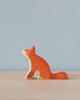 A Handmade Holzwald Sitting Fox figurine, categorized among sustainable toys, painted orange with a subtle smile, sitting on a flat surface against a soft blue background.