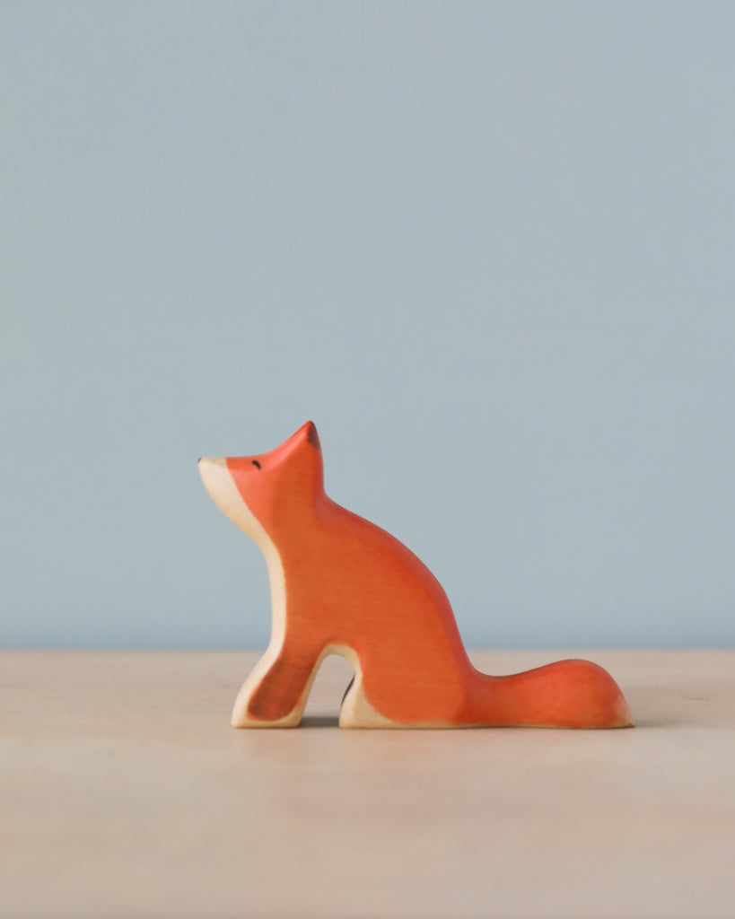 A Handmade Holzwald Sitting Fox figurine, categorized among sustainable toys, painted orange with a subtle smile, sitting on a flat surface against a soft blue background.