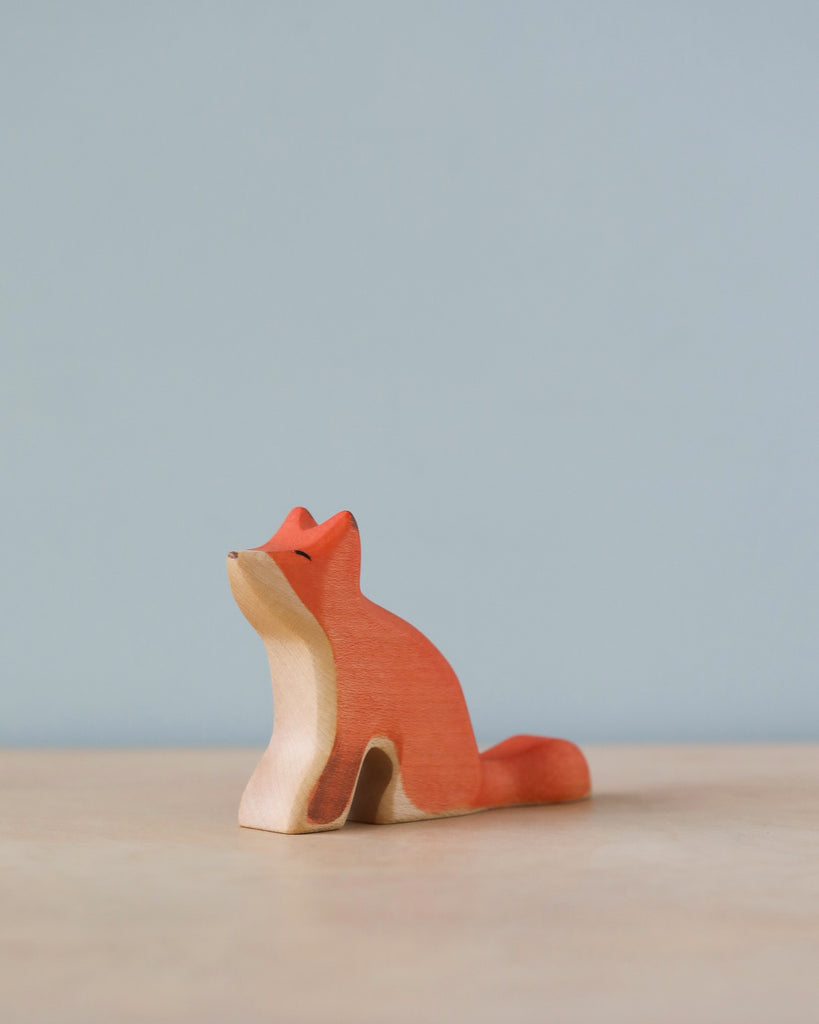 A Handmade Holzwald Sitting Fox painted in shades of orange and cream, sitting against a soft blue background, represents an artistic addition to any collection of sustainable toys.