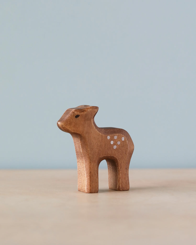 A Handmade Holzwald Standing Fawn, skillfully carved with visible wood grain and decorated with small white dots on its side, stands against a soft blue background.