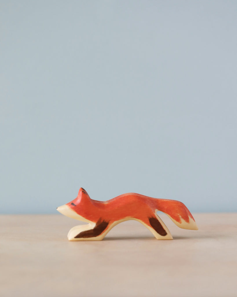 A Handmade Holzwald Small Fox figurine with white markings, positioned on a flat surface against a soft blue background. The fox is in a walking pose, painted in a simple, stylized manner.