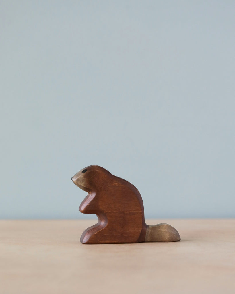 A high-quality Handmade Holzwald Beaver stands on a flat surface against a soft blue background. The figurine is simplistic with smooth contours and a natural wood finish.