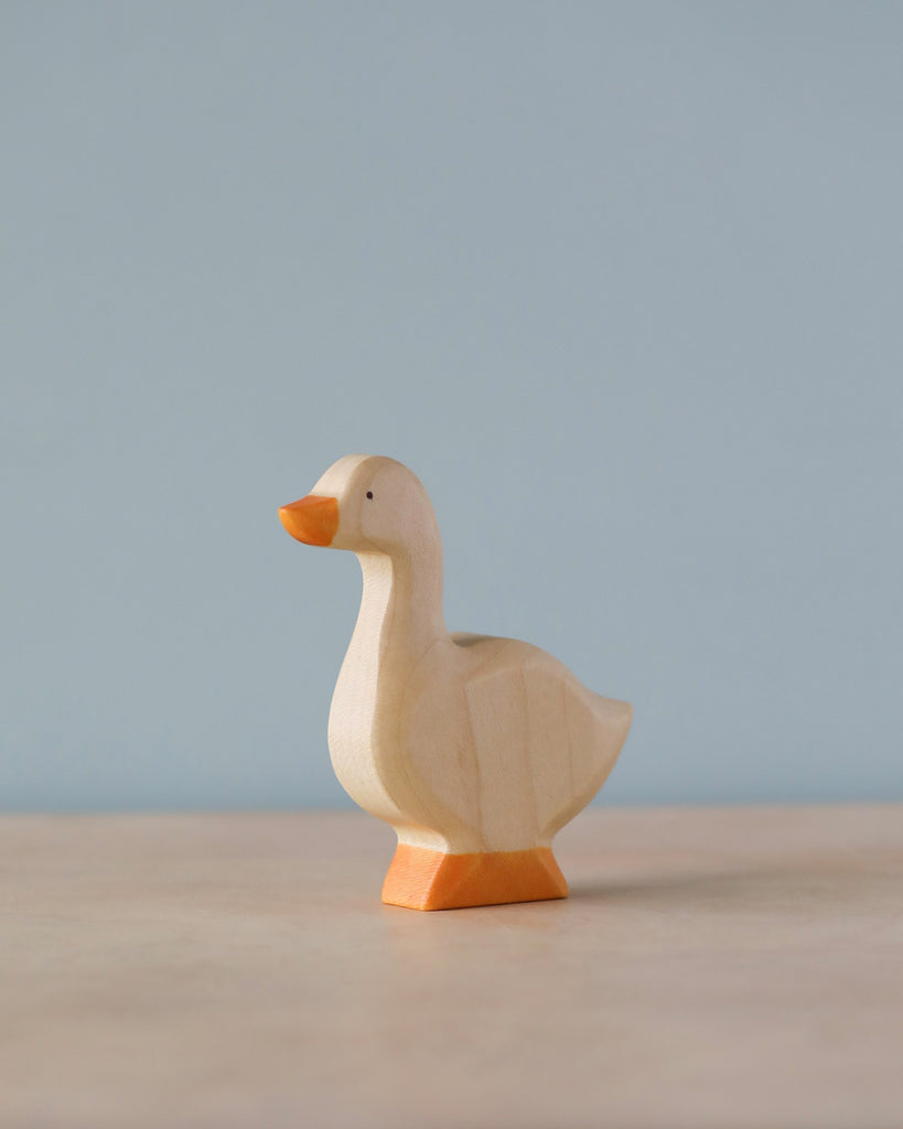 A sustainable Handmade Holzwald Goose figurine with a light body and orange beak and feet, standing against a soft blue background.