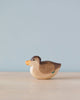 A Handmade Holzwald Swimming Duck figurine with a smooth finish, displayed against a soft blue background. The duck maintains a natural wood tone with a painted orange beak and small green detail.