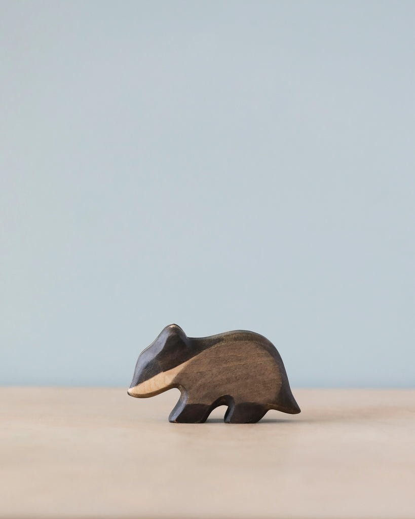 A Handmade Holzwald Badger figurine standing on a plain surface with a soft blue background. This sustainable toy highlights natural wood grains and a smooth finish.