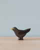 A small, dark wooden Handmade Holzwald Blackbird sculpture with a high-quality oil finish, featuring a simplistic design, standing on a flat surface against a light blue background.