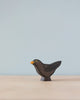 A Handmade Holzwald Blackbird, crafted from sustainable wood, with a black body and a yellow beak, standing on a flat surface against a plain light blue background.