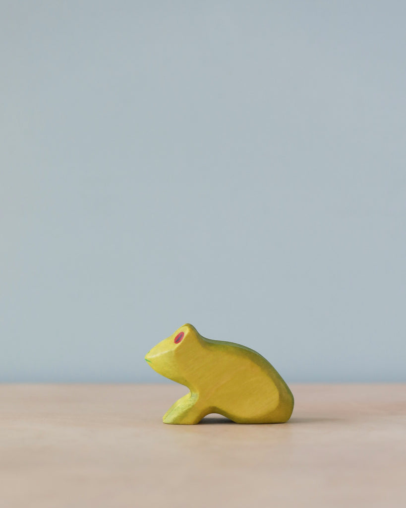 A simple Handmade Holzwald Frog toy painted in bright yellow with natural dyes and a red eye, placed against a soft blue background on a light surface. The frog is in a crouched position, facing.