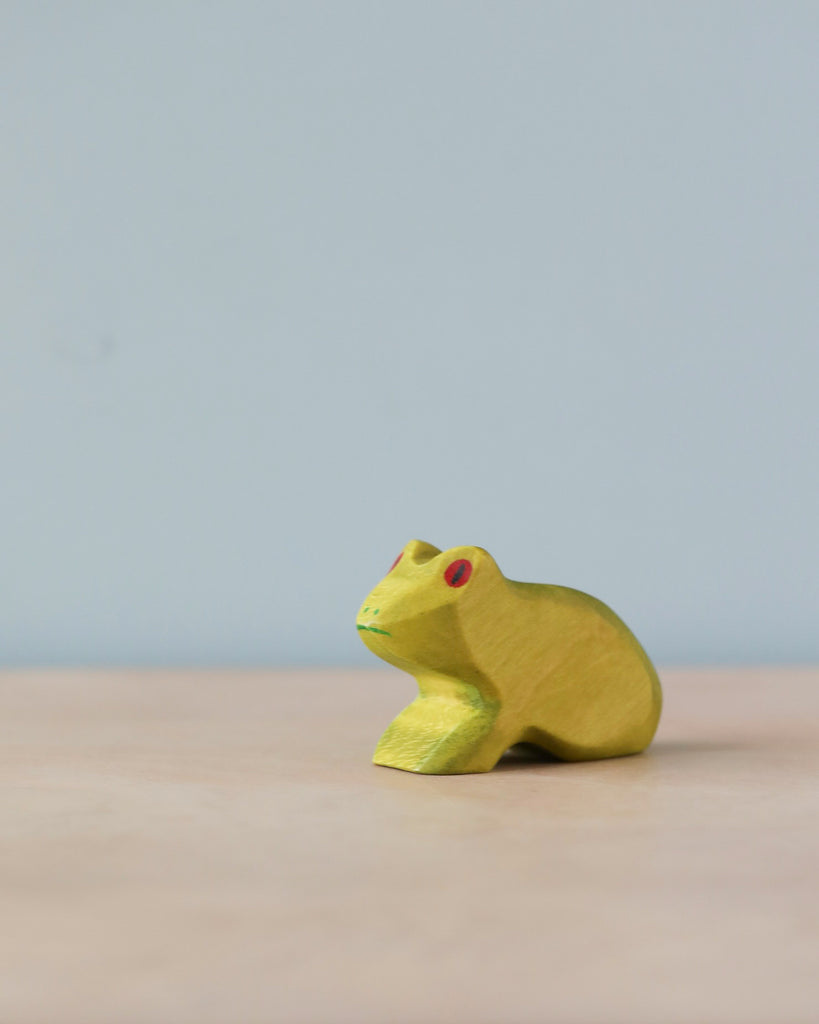 A small, handmade Holzwald Frog figurine painted with natural dyes yellow with red eyes, positioned on a light blue surface against a pale blue background.