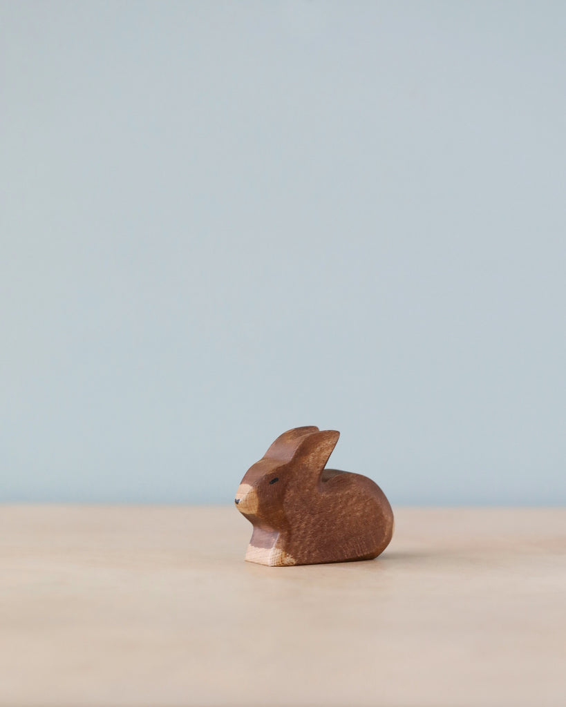 A Handmade Holzwald Small Brown Rabbit figurine set against a clear blue backdrop, positioned on a light beige surface. The rabbit is carved in profile, showing a simple, stylized form of sustainable toys.