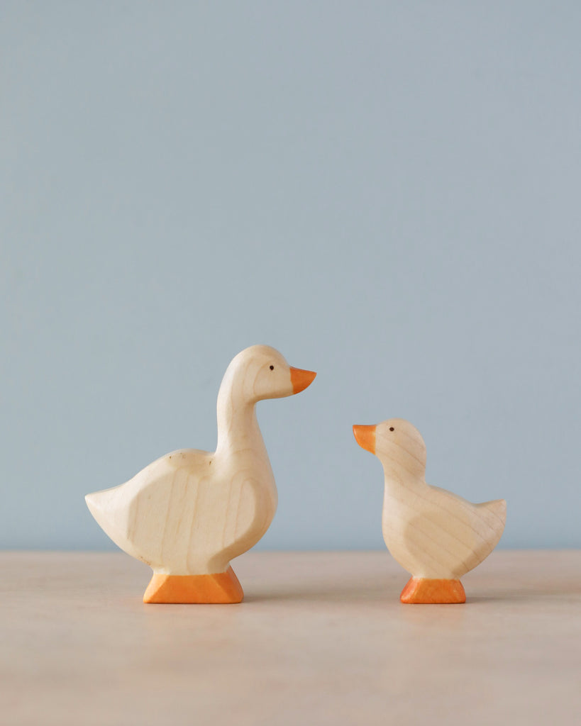 Two Handmade Holzwald Goose figurines, crafted with natural dyes, differing in size, facing each other on a plain surface against a soft blue background.