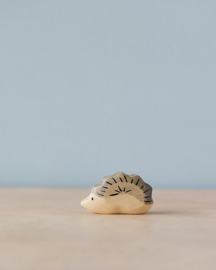 A small wooden figurine of a Handmade Holzwald Hedgehog Baby, featuring simple detailed carvings, sits on a smooth surface against a soft blue background. This piece is part of the Holzwald Brand's collection.